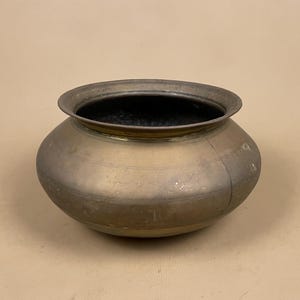 May include: A round, wide-mouthed brass vessel with a smooth, weathered patina. The bowl features a slightly flared rim and subtle horizontal etched lines encircling the midsection, showcasing a metallic gold and dark brown finish throughout.