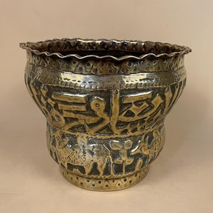 May include: A brass pot with a scalloped rim, featuring embossed Arabic calligraphy around the upper section and detailed engravings of camels and floral motifs on the lower body. The metal surface has a dark, aged patina with polished highlights.