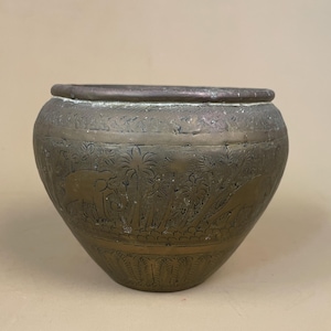 May include: A bronze-coloured, round pot with a wide rim. The pot features etched images of elephants, palm trees, and other foliage. The pot has a slightly tapered shape and sits on a neutral background.