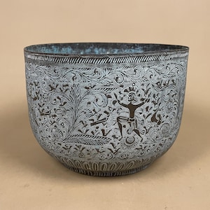 May include: A dark metal bowl featuring intricate white etched patterns of swirling floral vines and a central figure with arms raised. The interior and exterior surfaces show a weathered patina with hints of blue-green oxidation throughout.