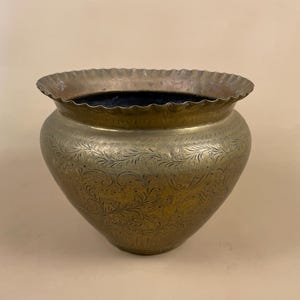 May include: A brass planter with an ornate, engraved floral design. The planter has a scalloped rim and a rounded, heart-shaped body.