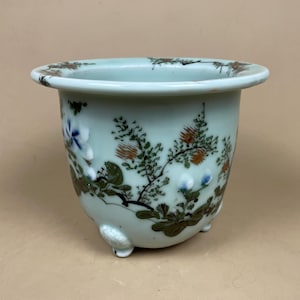 May include: A pale green ceramic plant pot with a wide rim and three small feet. It features hand-painted floral and botanical designs in blue, orange, and green. The pot has a rounded shape.