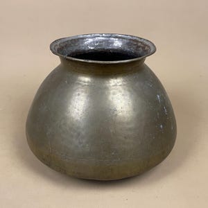 May include: A tarnished brass pot with a rounded body and a flared rim. The pot shows signs of age and wear, with a darkened interior. The exterior has a mottled appearance. This antique vessel is suitable for home decor, and is likely of Indian origin.