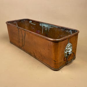 May include: A rectangular copper planter with a weathered patina and visible oxidation streaks on the interior walls. The exterior features a polished copper finish and a silver-toned metal lion head handle with a ring on the side panel.
