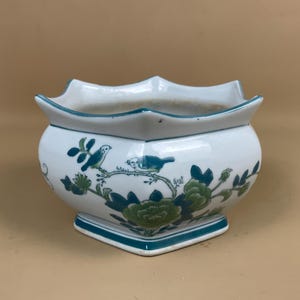 May include: White ceramic planter with a scalloped rim and a square base. The planter is decorated with teal and green floral and bird motifs. The rim and base are trimmed with teal. The planter has a rounded body.