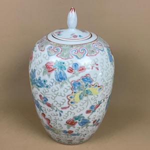 May include: A white ceramic jar with a lid, decorated with vibrant floral patterns in blue, red, and yellow. The jar has a rounded form and a small, white finial. The design includes a repeating pattern of the number 3.