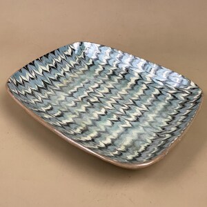 May include: Rectangular ceramic serving dish with a blue, white, and black chevron pattern. The dish has a slightly raised, light brown rim and rounded corners. The pattern creates a textured appearance.