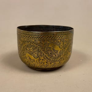 May include: A brass bowl with detailed etched patterns. The bowl has a dark interior and a gold exterior with repeating designs and animal figures. The overall color is a warm gold with dark accents.