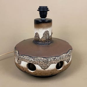 May include: A ceramic table lamp base featuring a wide, rounded body with a brown and cream glaze. The surface has a textured, speckled finish with circular cutouts around the lower section and a black plastic light socket at the top centre.