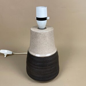 May include: A ceramic table lamp with a speckled beige upper section and a textured dark brown lower section. The lamp has a white bulb socket with a black band and a white power cord with an on/off switch.