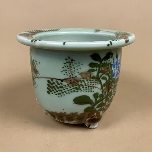 May include: A small, pale green ceramic planter with a rounded shape and three small feet. It features hand-painted brown, green, and blue floral and landscape designs. The rim has a brown trim.