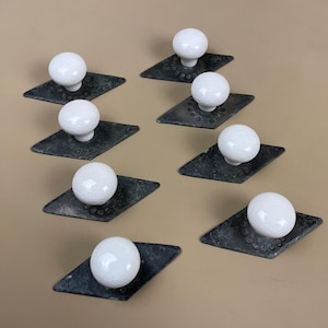 May include: Eight cabinet knobs with white ceramic spherical handles on dark, diamond-shaped metal backplates. The backplates have a textured surface. The knobs are arranged in a pattern against a neutral background.