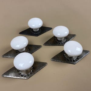 May include: Five cabinet knobs. Each has a white, crackled ceramic sphere on a diamond-shaped, hammered metal base. The metal bases have a dark, aged finish with small, circular indentations around the knob's stem. The knobs are for cabinets.