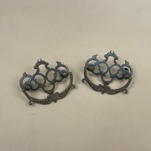 May include: Two antique metal drawer pulls with intricate, ornate designs. The pulls are in a silver finish and have a decorative, scroll-like pattern.