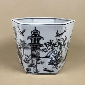 May include: A white, hexagonal planter with a black Chinoiserie design. The design features a pagoda, birds, trees, and figures. The planter has a black rim and a neutral background.
