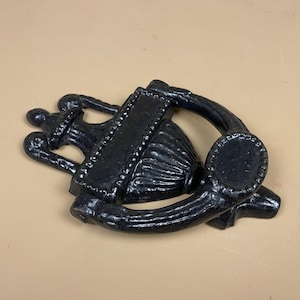 May include: Black cast iron door knocker with an ornate design. It features a rectangular plate, a bell-shaped element, and a curved handle. The design is vintage-inspired and suitable for a front door.