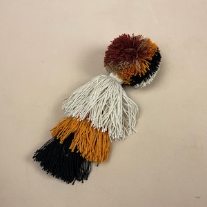 May include: A multi-coloured tassel with a round pom-pom at the top. The tassel is made of white, black, orange, and brown yarn.
