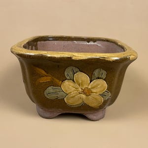 May include: A square, brown ceramic planter with a yellow flower design. The planter has a light brown rim and three small feet. The interior is a muted purple colour. The flower has a detailed, raised design with green leaves.
