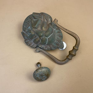 May include: A vintage brass door knocker with a detailed face design. The knocker is attached to a brass ring and has a separate brass knob.
