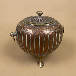 May include: A round copper vessel with a matching lid, featuring vertical fluted panels and a decorative engraved band near the top. The container stands on three brass feet and includes two side ring handles, showing a natural aged patina.