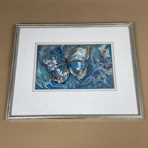 May include: A framed watercolour painting of a butterfly. The butterfly has blue and white wings with intricate details. The background is a textured blue with floral accents. The frame is silver.