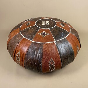 May include: Circular leather ottoman in brown and dark brown tones, featuring white stitching and diamond and geometric patterns. The ottoman has a central emblem and is designed in a circular shape.