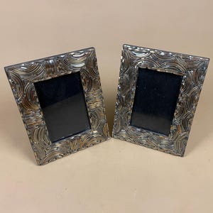 May include: A pair of decorative photo frames with a mosaic design in silver and gold tones. Each frame features a black interior and a rectangular shape. The frames are displayed against a neutral background, suitable for displaying photographs.