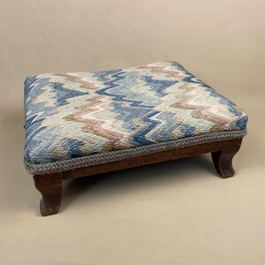 May include: A small, square footstool with a patterned fabric top in shades of blue, pink, and beige. The fabric has a zig-zag design. The footstool has a dark wooden frame and four curved legs.