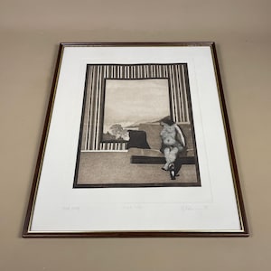 May include: Framed monochrome print showing a figure seated on a sofa, looking out of a window. The artwork has a minimalist style with vertical lines and a landscape view. The frame is a dark brown colour.