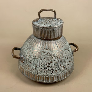 May include: A round metal container with a lid and two side handles. The surface features an intricate engraved floral pattern with a dark copper finish and a light, weathered patina filling the recessed areas of the detailed metalwork.