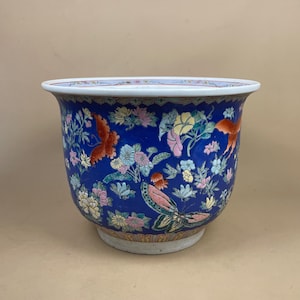 May include: A deep blue ceramic planter featuring a vibrant, multi-coloured pattern of various flowers and butterflies. The bowl-shaped vessel has a wide, flared white rim and a tapered base, decorated with intricate floral motifs in pink, yellow, and green.
