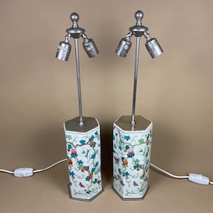 May include: Two decorative table lamps. Each lamp has a hexagonal base decorated with a floral and butterfly pattern in green, orange, and pink. The lamps have a silver-coloured metal stem and three light sockets. The lamps are connected to a power source.