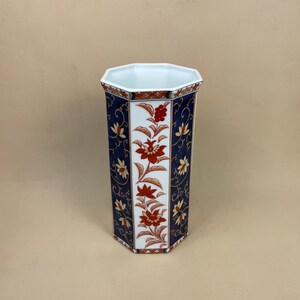 May include: A white porcelain vase with an octagonal shape and a blue and gold floral design. The vase features red flowers and leaves.