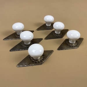 May include: Seven white ceramic cabinet knobs with a round shape are mounted on diamond-shaped, aged metal backplates. The backplates have a hammered texture and are arranged on a neutral background. These knobs are suitable for furniture restoration or DIY projects.