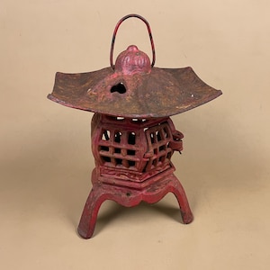 May include: A red cast iron Japanese garden lantern. The lantern has a square base with three legs, a latticed body, and a flared top with a handle. The lantern is weathered with rust and has a hole in the top.