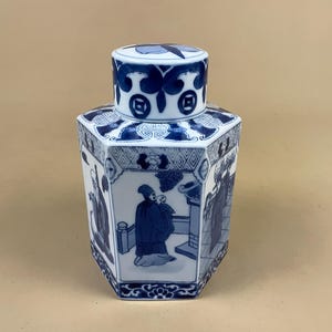 May include: A hexagonal ceramic jar with a lid, featuring blue and white hand-painted illustrations of figures in traditional attire. The body displays detailed scenes within panels, while the lid is decorated with a blue floral motif.