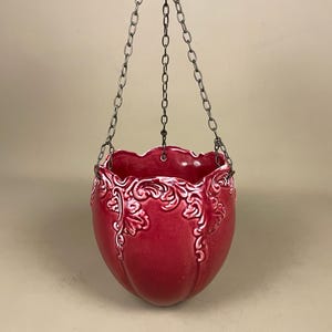 May include: A red ceramic hanging planter with a white floral design. The planter is suspended by a silver chain. The rim has a scalloped edge. A stylish addition for indoor or outdoor plants. (approx. 20cm diameter)