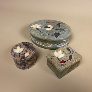 May include: Three decorative stone boxes. The boxes are oval, round, and square, with floral inlay designs in cream, black, and brown. The oval box is green with a cream and black floral design. The round box is brown with a cream floral design.