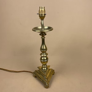 May include: A vintage brass table lamp with an ornate design. The lamp has a triangular base, a slender stem with a circular dish, and a bulb socket. The lamp is made of brass and has a warm, golden colour. The lamp's height is approximately 30 cm.