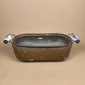 May include: An antique copper rectangular tub with two handles. The handles are white ceramic with blue floral designs. The copper has a patina, showing signs of age. The tub is empty and sits on a neutral background.