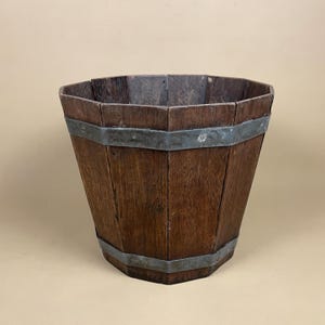 May include: A weathered wooden bucket, featuring a dark brown hue and a tapered design. The bucket is held together by two metal bands. The wooden staves are arranged vertically, giving it a rustic, aged look. The bucket's height is approximately 30 cm.