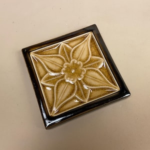 May include: A square ceramic tile with a brown border and a raised, beige floral design. The flower has five petals and a centre with a smaller flower.