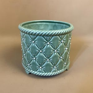 May include: A pale green ceramic plant pot with a quilted design and white embellishments. It features a rope-style trim at the top and base. The pot is cylindrical and rests on three small feet.