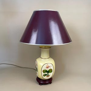 May include: A table lamp with a maroon lampshade and a cream-coloured base. The base features hand-painted floral designs and sits on a dark brown wooden stand. The lamp has a silver trim around the lampshade.