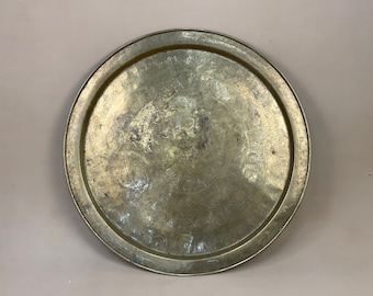 May include: A round, brass serving tray with an ornate, engraved design. The tray has a raised rim and a slightly domed center.