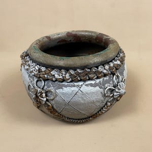 May include: A small, round, silver planter with a decorative braided rope trim and ornate metal accents. The planter has a distressed, aged look.