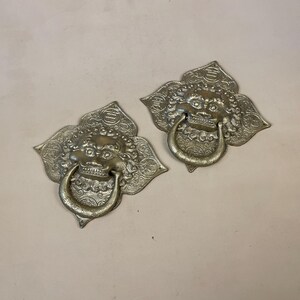 May include: Two ornate brass door knockers shaped like a lion's head with a ring handle. The knockers are detailed with intricate floral patterns and have a gold finish.