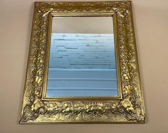 May include: A rectangular mirror with a gold-colored, ornate frame. The frame features detailed floral and foliate relief carvings. The mirror reflects a light-colored wall and a glimpse of a white brick wall.