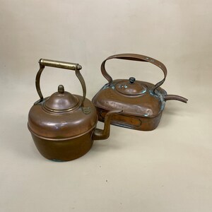 May include: Two antique copper tea kettles. One kettle has a handle and spout, the other has a handle and a long spout. Both kettles have a tarnished finish.