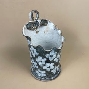 May include: A ceramic utensil holder with a white interior and a dark teal exterior decorated with white floral patterns. The top edge is scalloped, and it has a loop handle. The holder is approximately 15 cm tall.